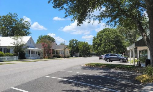 Paved small-town Florida street and parking area beside trees and modest historic low-rise buildings.